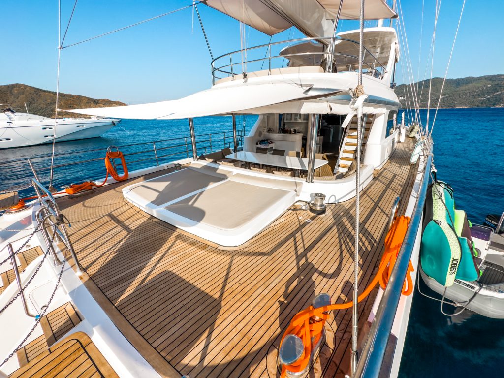 A luxury yacht for charter with a spacious wooden deck, shaded lounge area, and comfortable seating is anchored in calm blue water near distant hills. Inflatable water toys hang on the side under a clear sky.