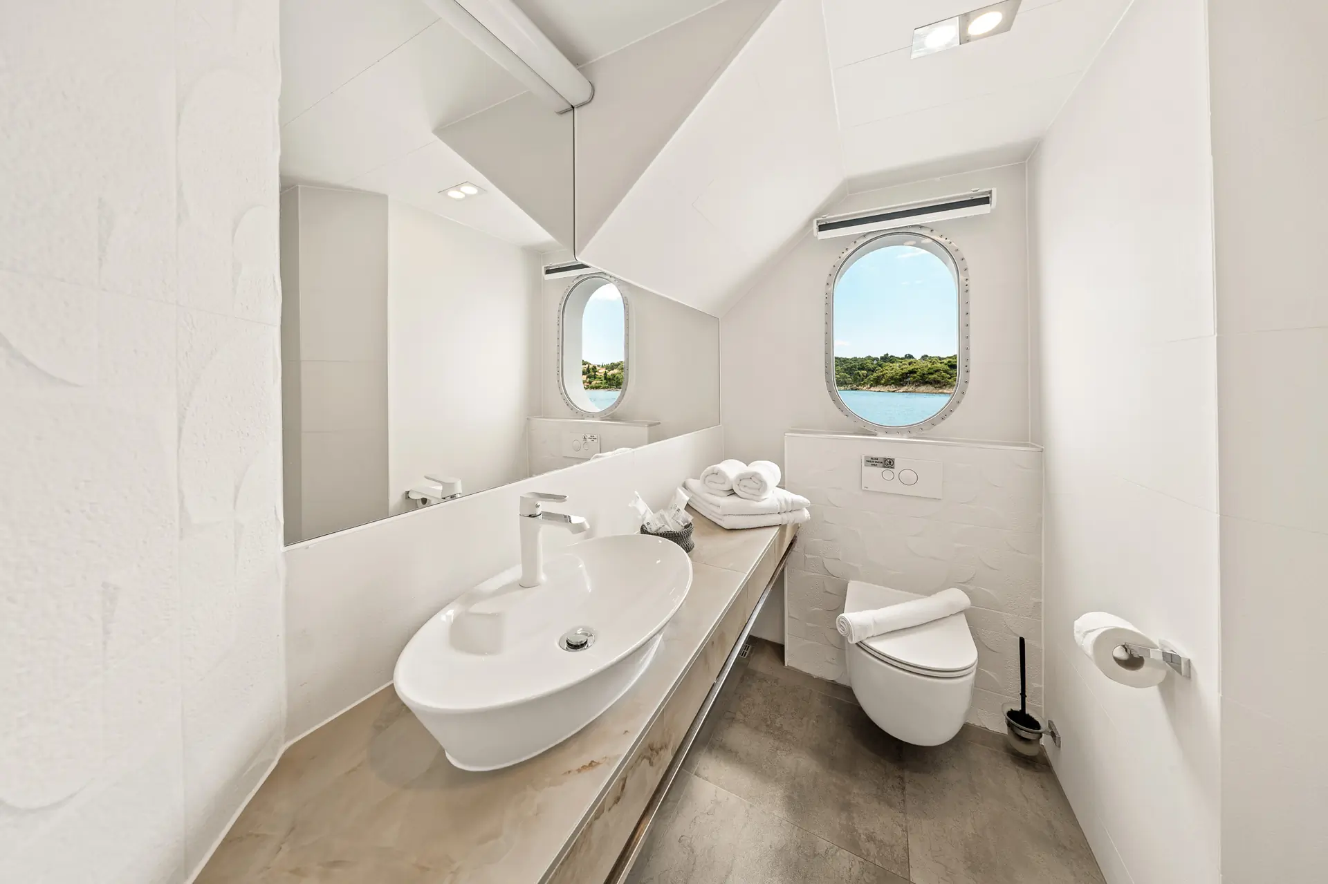 Modern white bathroom with a round window, oval sink on a long countertop, wall-mounted toilet, folded towels, and a large mirror reflecting natural light and an outdoor view.