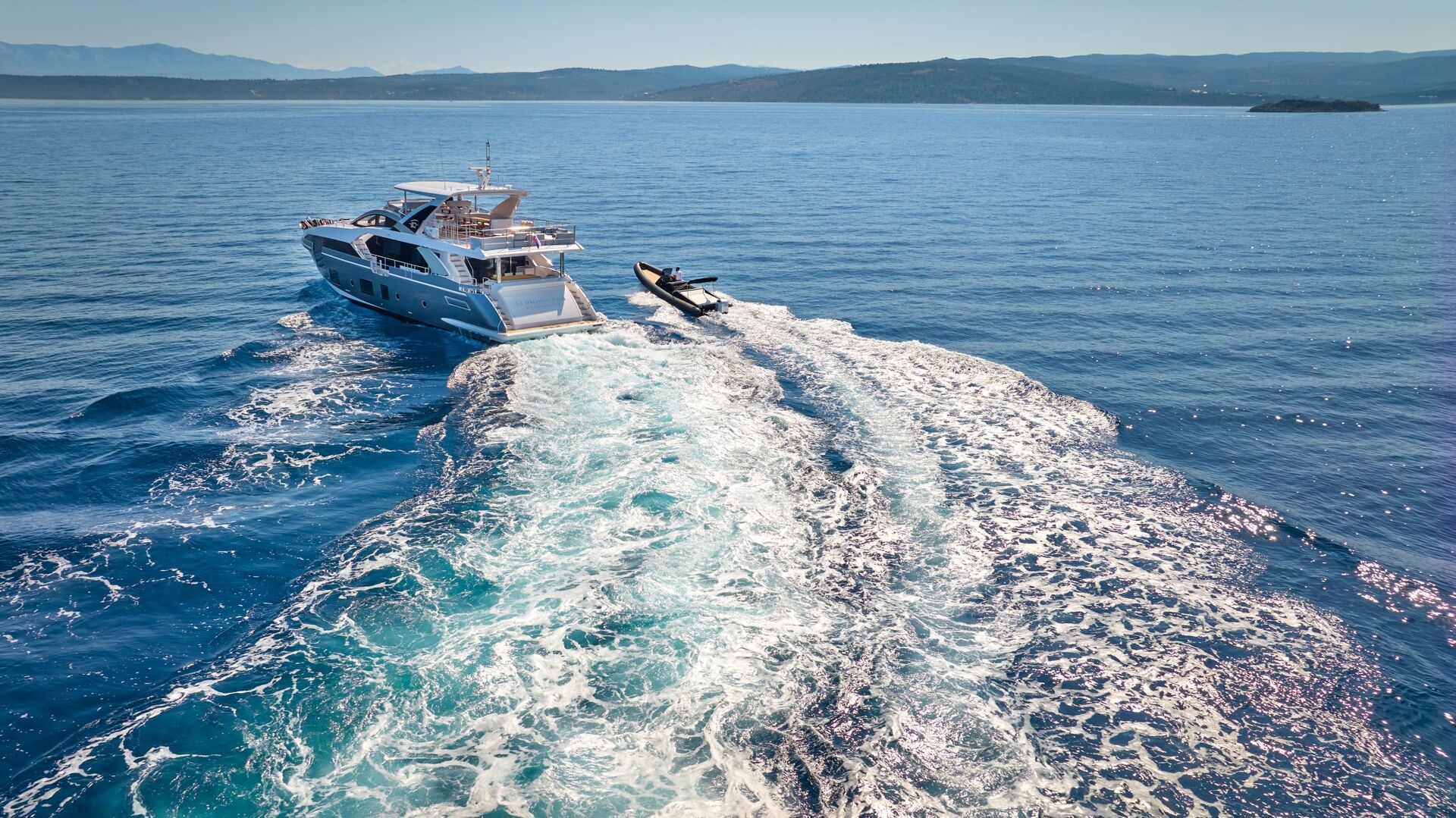 A white yacht travels through clear blue water, leaving a foamy wake behind as it tows a small boat. The distant shoreline and mountains are visible under a clear sky.