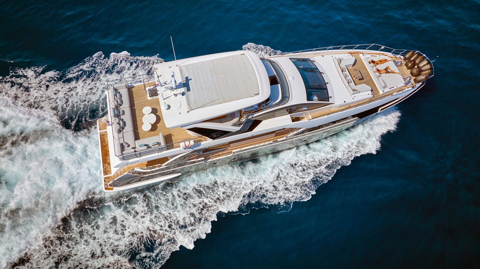 Aerial view of a luxury white yacht cruising through deep blue water, leaving a foamy wake behind it. The yacht has sunbathing areas, a dining table, and a modern design.