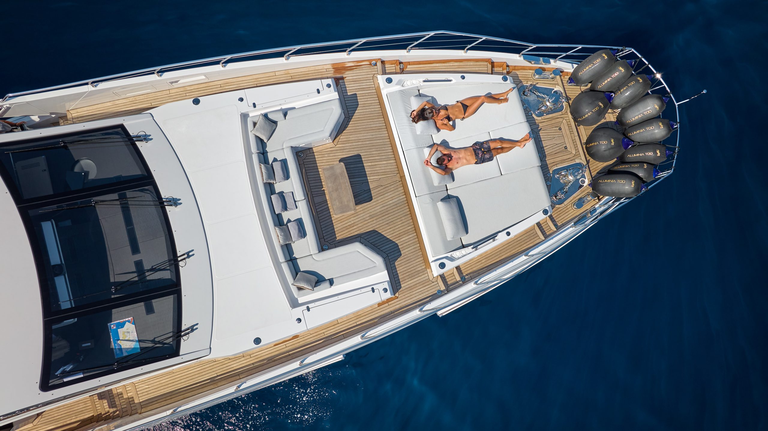 A top-down view of two people sunbathing on the deck of a luxury yacht, surrounded by blue ocean water. The yachts white and wooden finishes and multiple engines are visible.