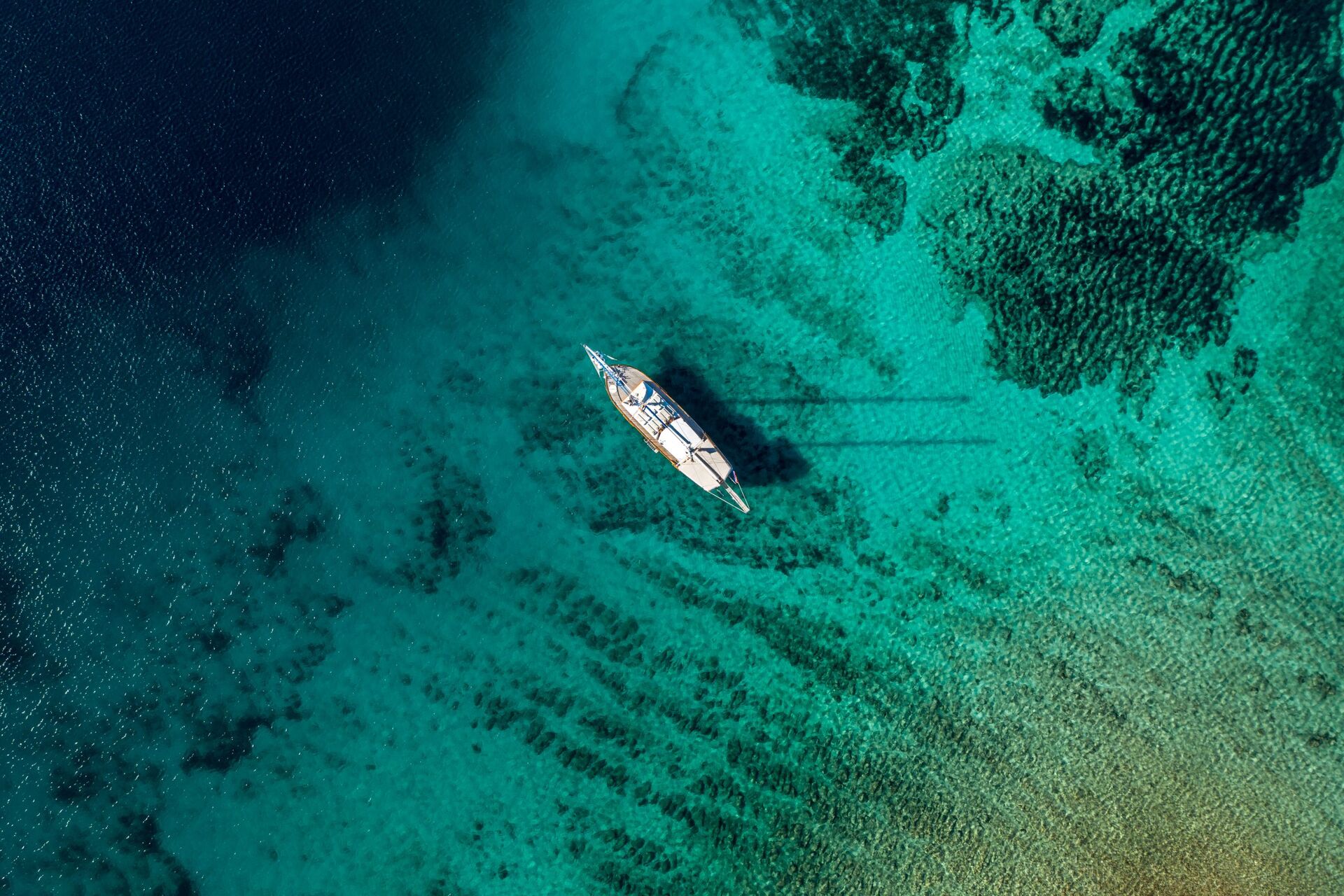Aerial view of a sailboat floating on clear turquoise water, with dark patches of underwater vegetation visible below the surface.