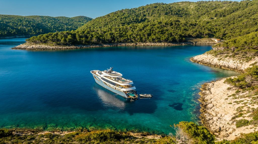 A large white yacht is anchored in a clear blue bay surrounded by forested hills and rocky shores under a sunny sky.