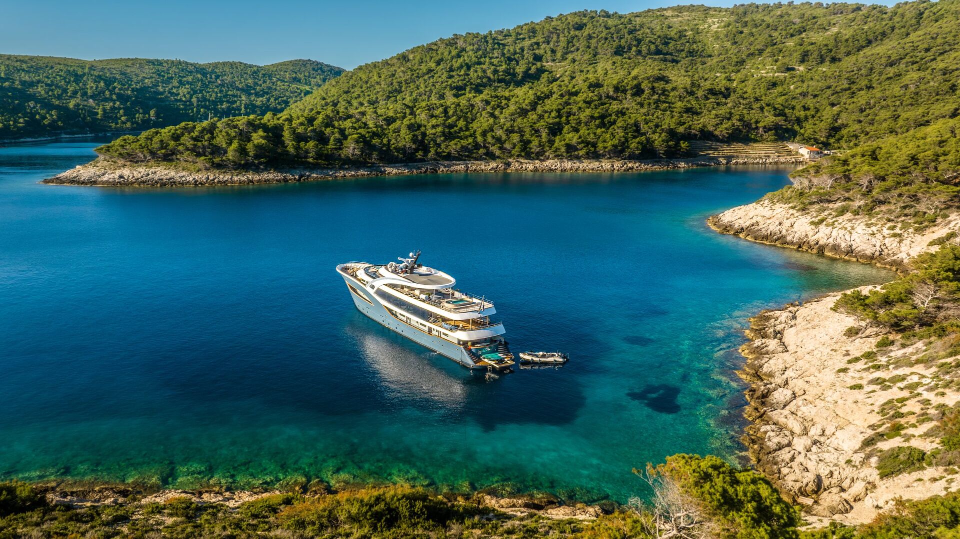 A large white yacht is anchored in a clear blue bay surrounded by forested hills and rocky shores under a sunny sky.