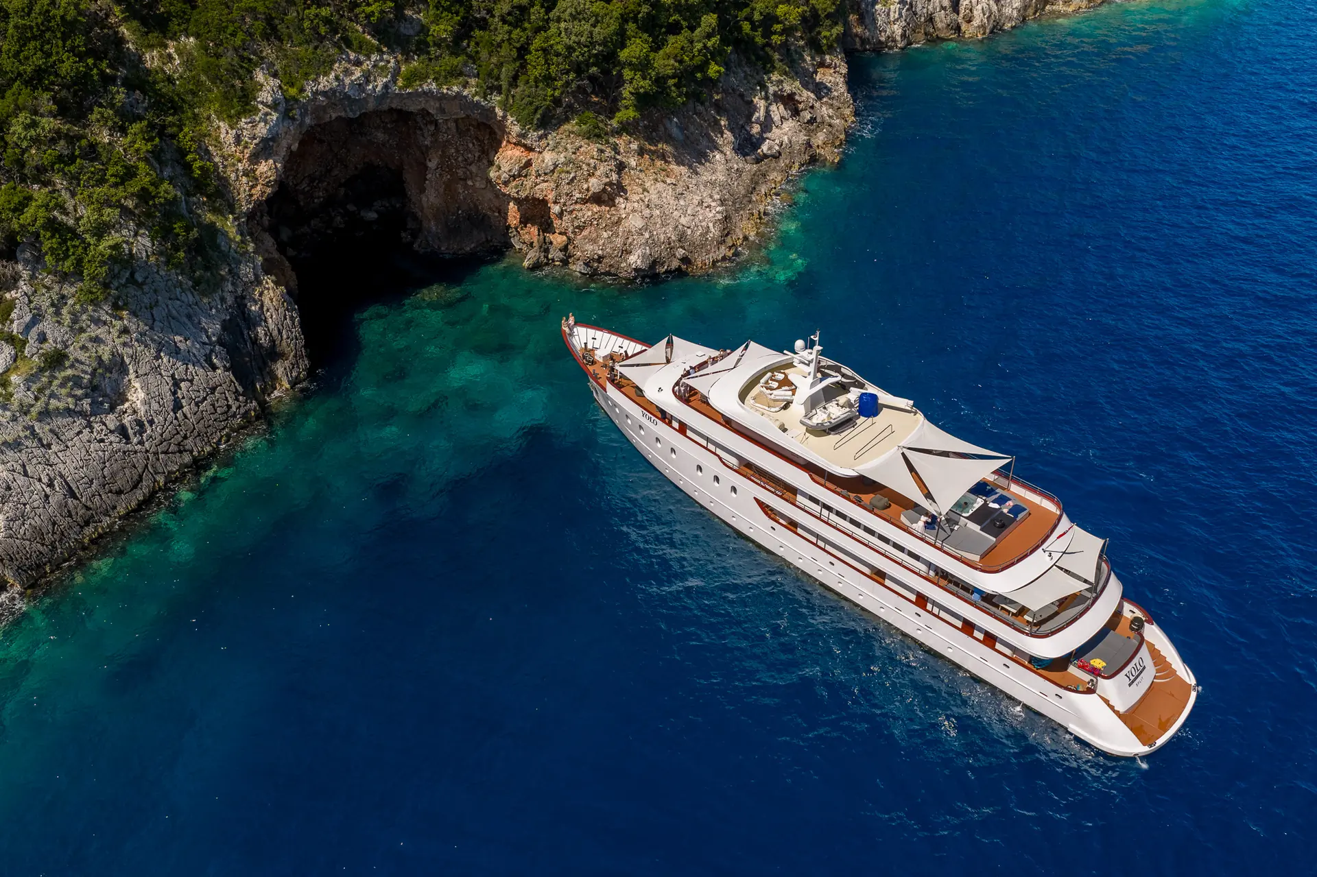 A large white yacht floats on clear blue water near a rocky coastline with lush green trees and a cave at the waters edge.