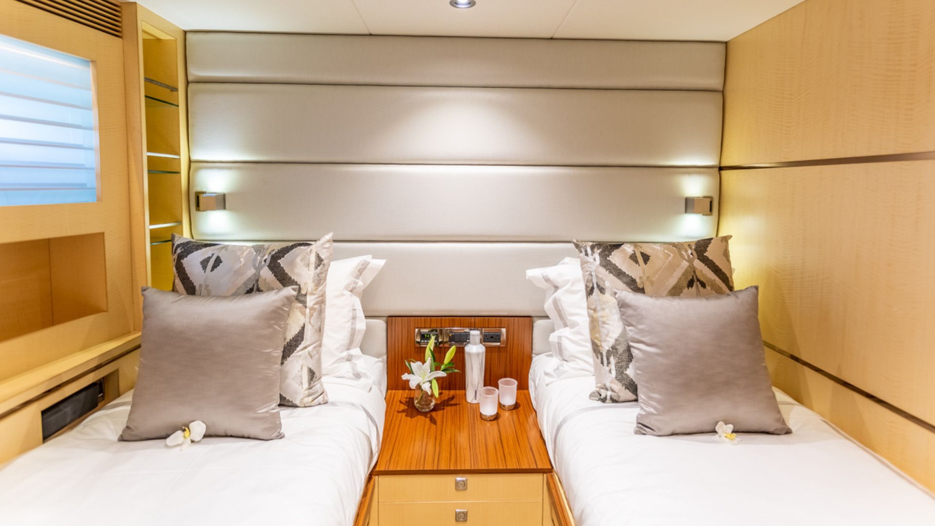 A modern yacht cabin with two single beds, each with patterned pillows and gray cushions, separated by a wooden nightstand with flowers and glasses; light wood paneling and soft wall lights create a cozy atmosphere.