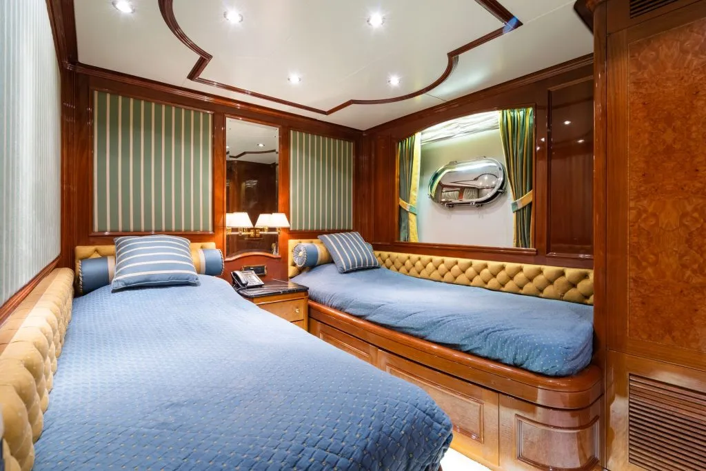 Luxurious yacht cabin with two single beds, blue bedding, striped pillows, wood paneling, wall lamps, a telephone on a bedside table, striped curtains, and a round porthole window.