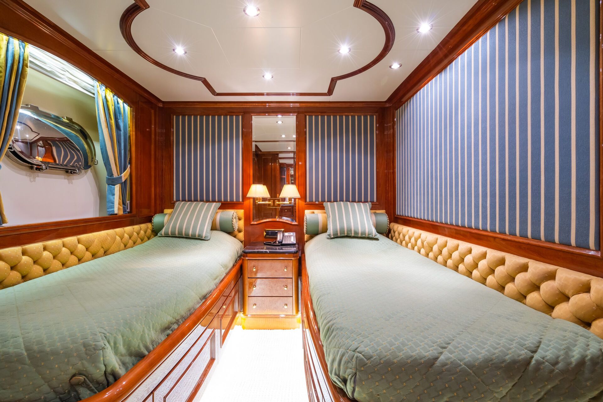 A luxurious cabin with two single beds, tufted leather headboards, striped pillows, a central nightstand with lamps, wood paneling, striped blue wallpaper, and a large mirror at the head of the beds.
