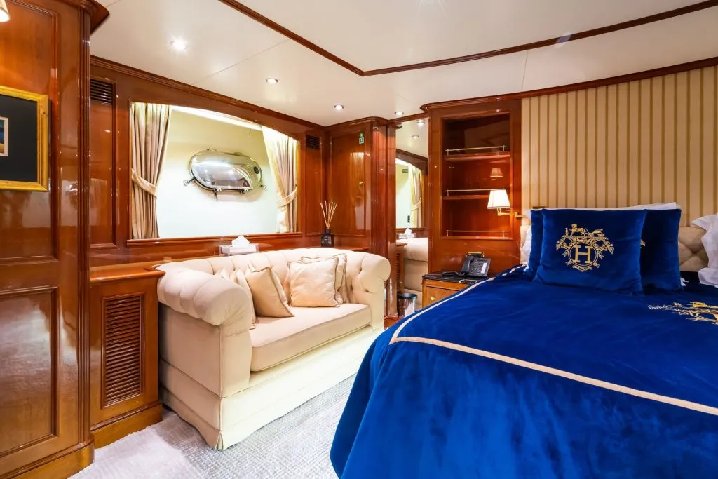 Luxurious yacht cabin with polished wood walls, a cream-colored sofa with cushions, and a blue bedspread featuring a gold emblem. Soft lighting, built-in shelves, and a mirrored window create an elegant, cozy atmosphere.