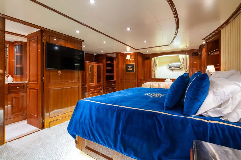 Luxurious bedroom with wooden walls and furniture, a large bed with blue bedding and pillows, a wall-mounted TV, and a well-lit, elegant interior. A mirror and open doorway add depth to the room.