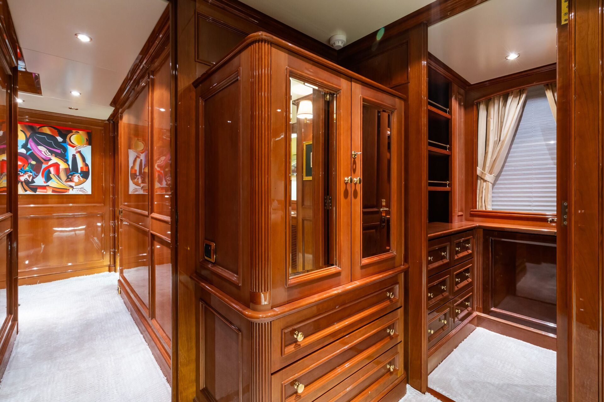 Luxurious wood-paneled walk-in closet with ample drawers, cabinets, a window with beige curtains, carpeted floor, and abstract artwork on the wall. The space is well-lit with recessed ceiling lights.