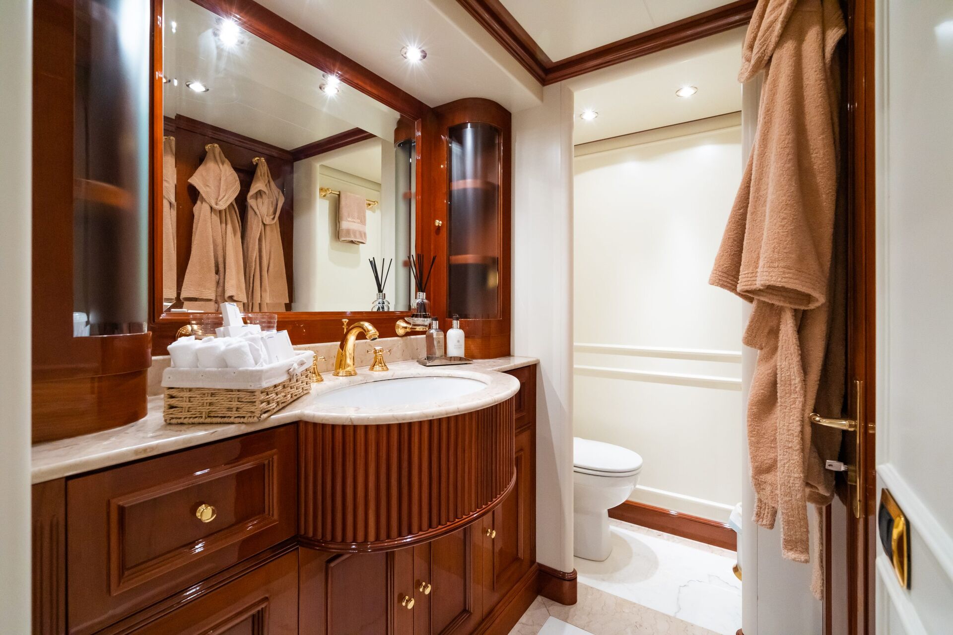 Luxurious bathroom with wooden cabinetry, marble countertop, gold faucets, and large mirror. Towels and robes hang on the walls, and a basket with toiletries sits beside the sink. Toilet visible in the corner.