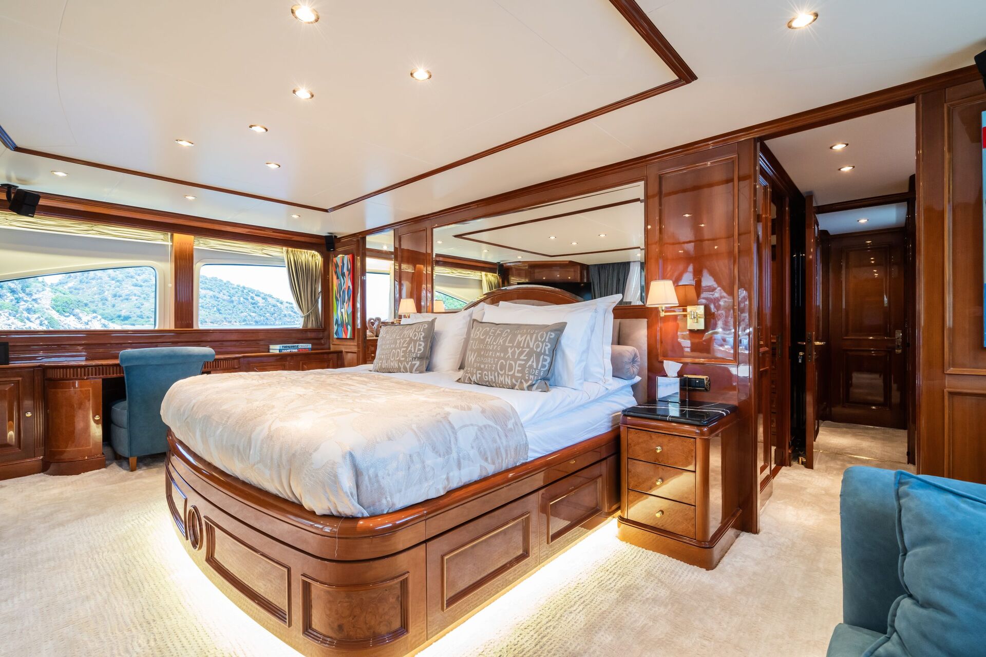 A luxurious yacht bedroom with a large bed, polished wood paneling, plush bedding, large windows with ocean views, a desk with a chair, and soft ambient lighting creating an elegant, inviting atmosphere.