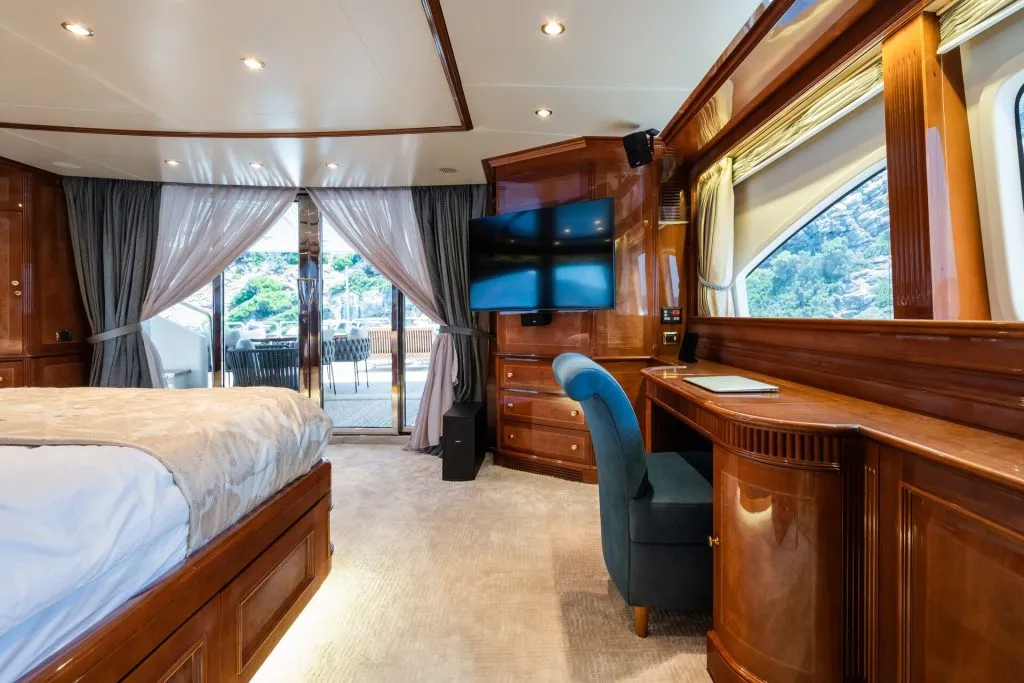 Luxurious yacht cabin with a large bed, polished wood furniture, blue chair at a desk, wall-mounted TV, and glass doors opening to an outdoor deck with scenic views. Natural light fills the spacious room.