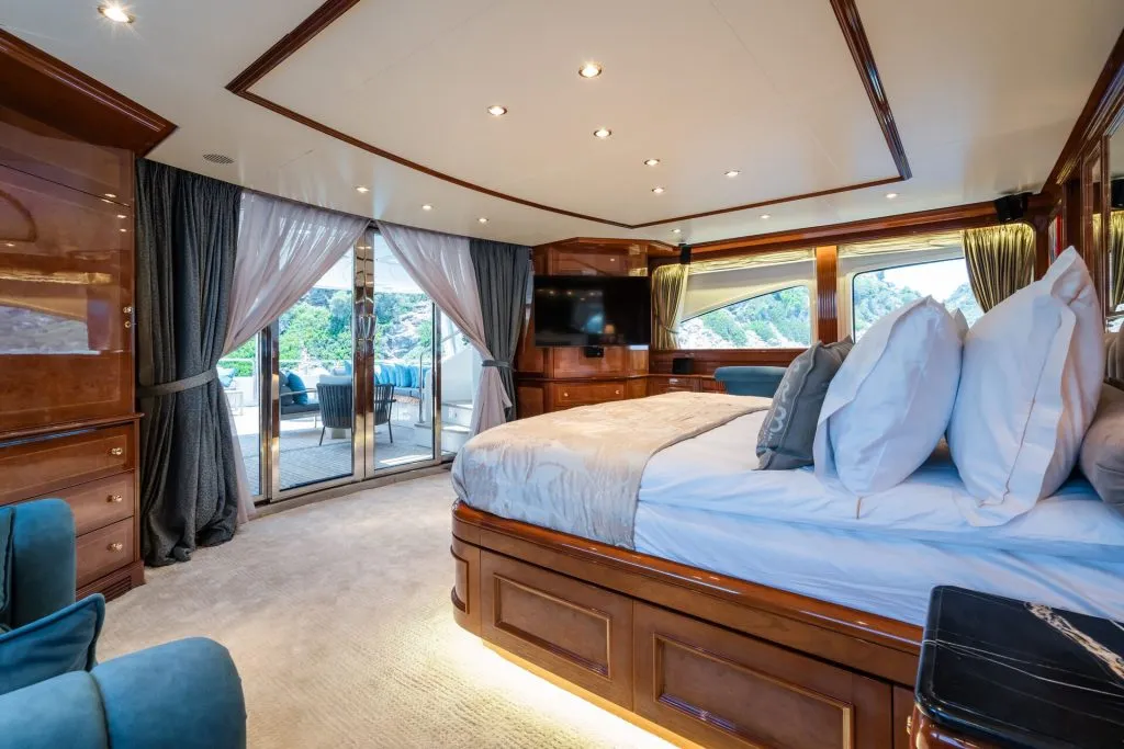 Luxurious yacht bedroom with a large bed, plush pillows, wooden furnishings, a wall-mounted TV, large windows with scenic views, and glass doors leading to an outdoor seating area. Soft lighting creates a cozy atmosphere.