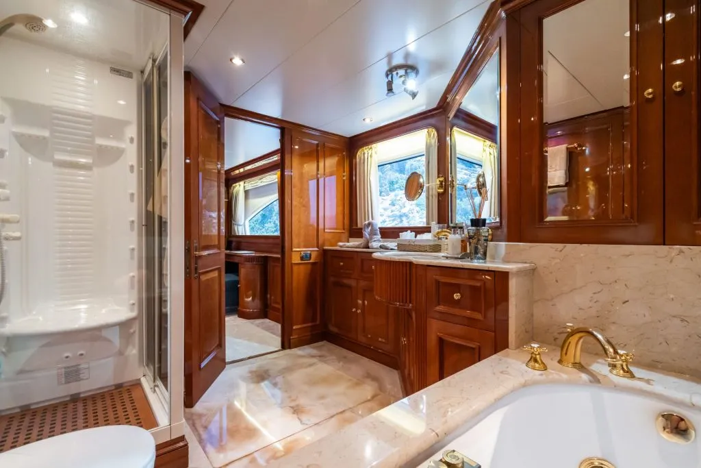 Luxurious bathroom with wooden cabinets, marble countertop, gold fixtures, a large bathtub, a glass-enclosed shower, and a window with yellow curtains, reflecting an elegant and spacious design.