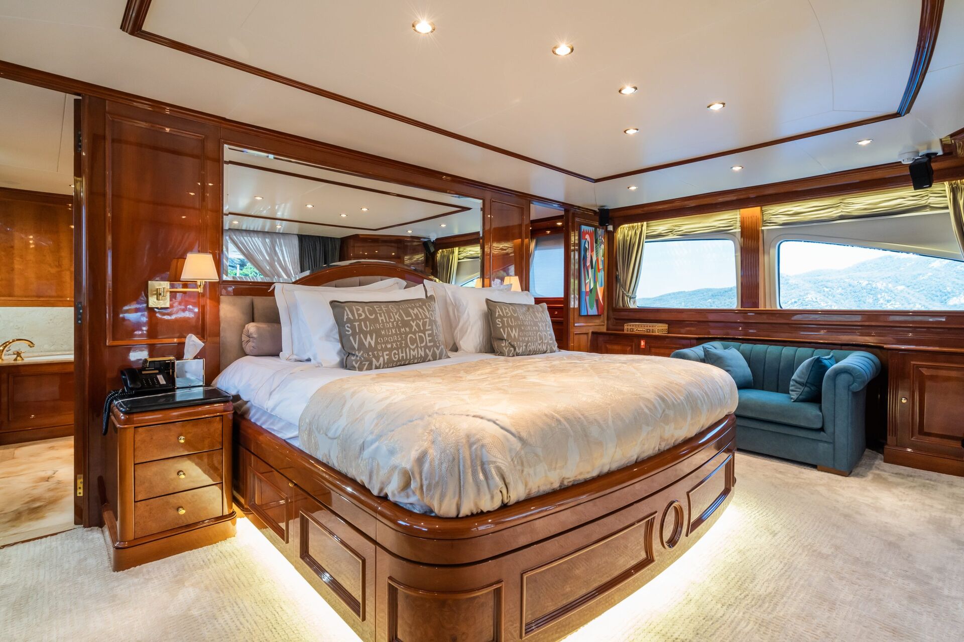 Luxurious yacht bedroom with wood-paneled walls, a large bed with gray pillows, bedside tables, mirrored wall, green velvet sofa, and large windows offering scenic views of mountains and water.
