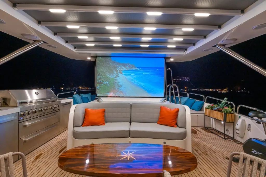 A luxurious yacht deck at night features a large screen showing a beach scene, a modern grill, a white sofa with orange pillows, a wooden table, and city lights visible in the background.