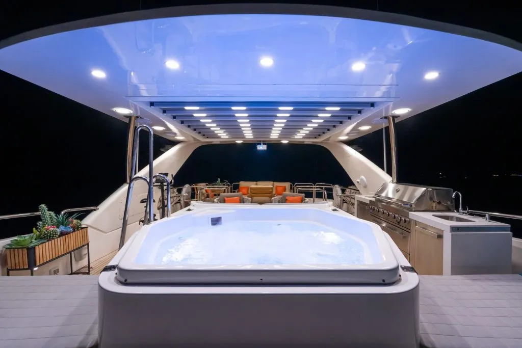 A luxury yacht deck at night features a lit hot tub, outdoor lounge seating with orange cushions, a grill, and a fruit basket, all under a modern illuminated canopy.