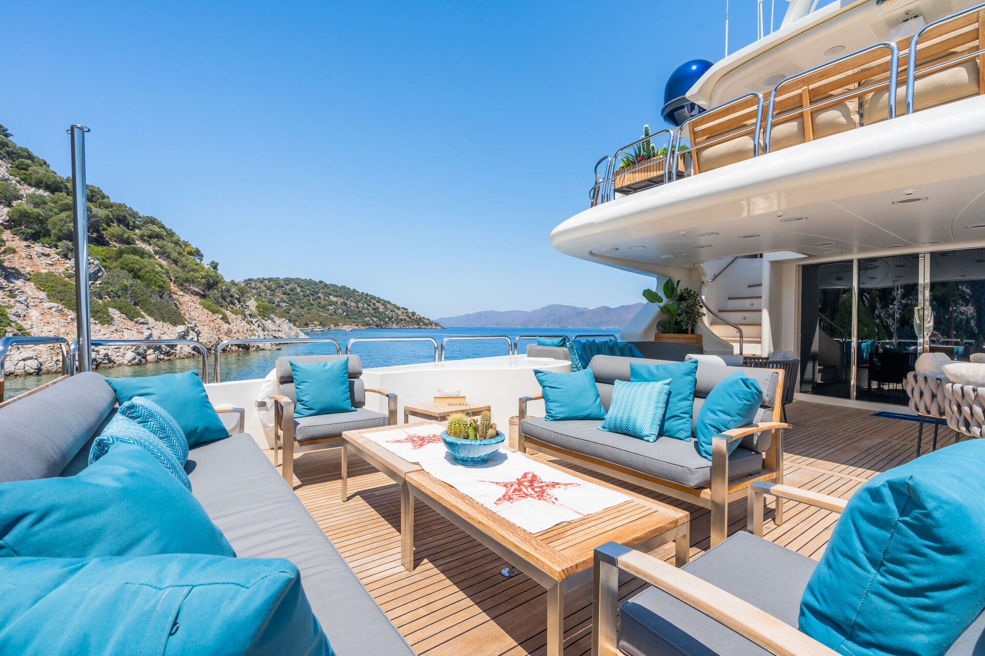 A spacious yacht deck with cushioned seating, blue and white pillows, a wooden table with decorative items, and ocean views with hills in the background under a clear sky.