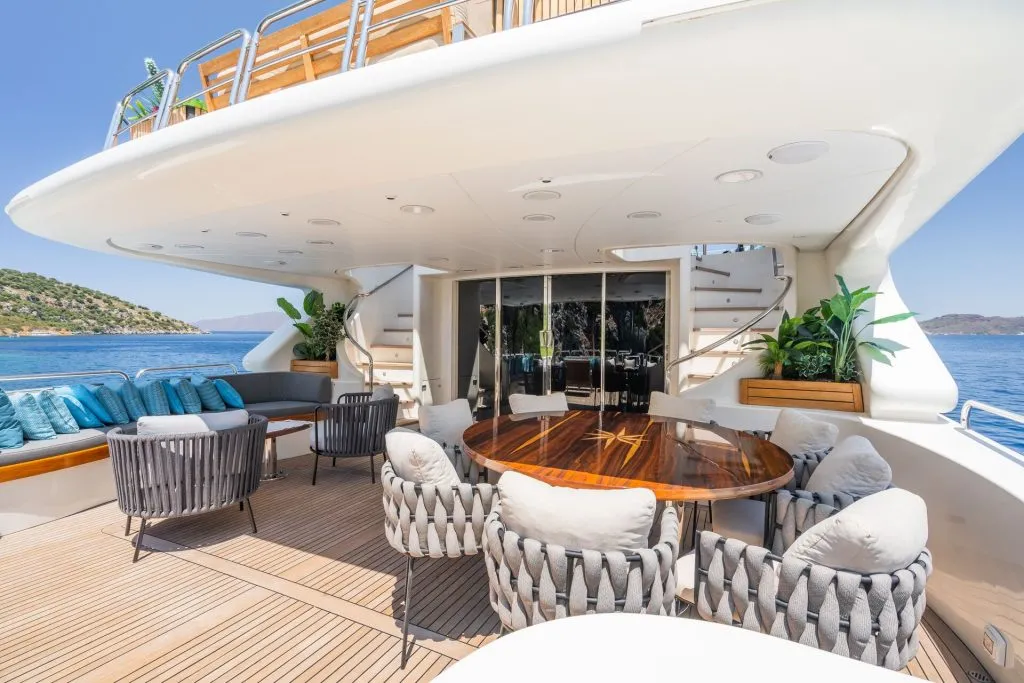 A luxurious yacht deck with a polished wooden dining table, cushioned chairs, lounge seating, potted plants, and views of blue sea and green hills under a clear sky.