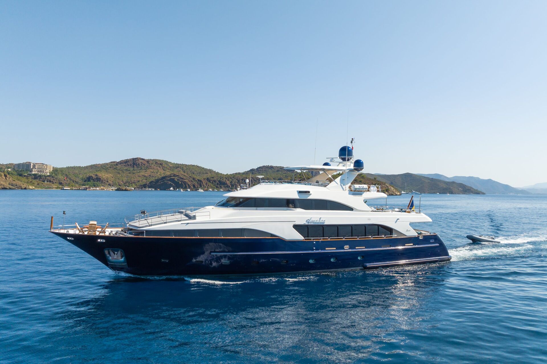 A luxury motor yacht with a white upper deck and dark hull sails on calm blue water, with green hills and clear sky in the background.