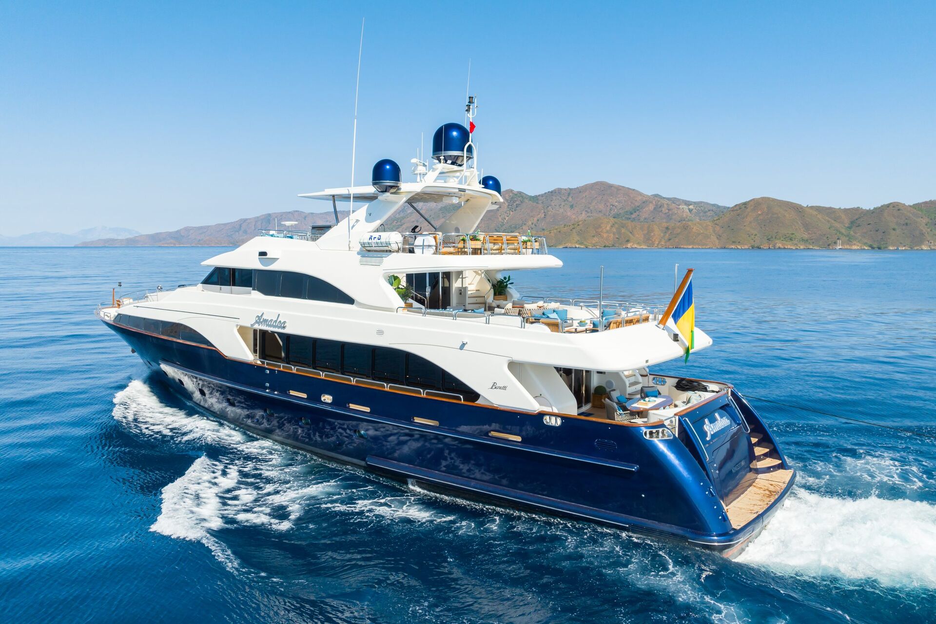 A large luxury yacht with a blue and white exterior sails on calm blue water near a mountainous coastline under a clear sky. The yacht has multiple decks and a flag at the stern.