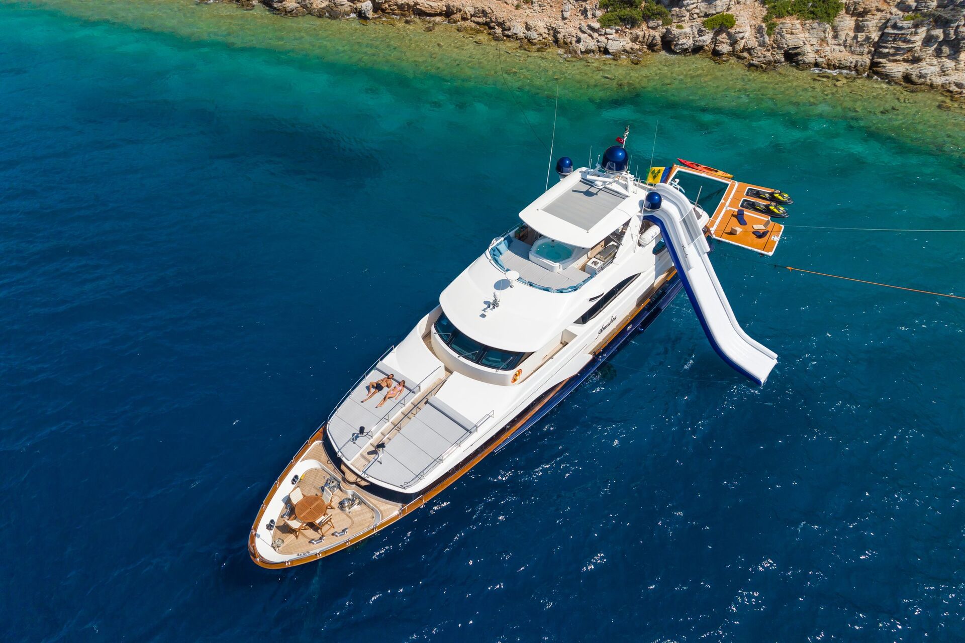 Aerial view of a white luxury yacht anchored in clear blue water near a rocky shoreline, with a water slide attached to the upper deck and floating platforms in the sea nearby.