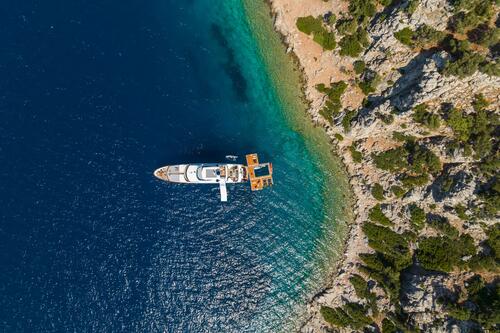 A yacht anchored near a rocky, tree-covered coastline, with clear blue and turquoise water contrasting against the shore. Small platforms and a dinghy float beside the yacht.