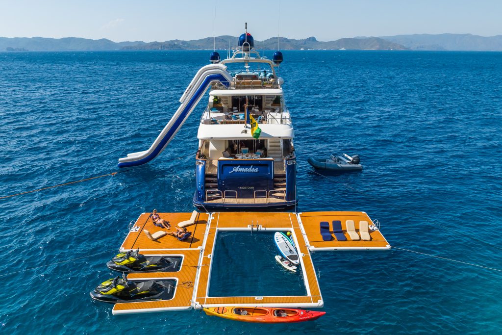A luxury yacht anchored at sea with a large slide, floating platforms, jet skis, kayak, and lounge chairs. People are sunbathing and swimming in a netted pool on the ocean. Mountains are visible in the background.