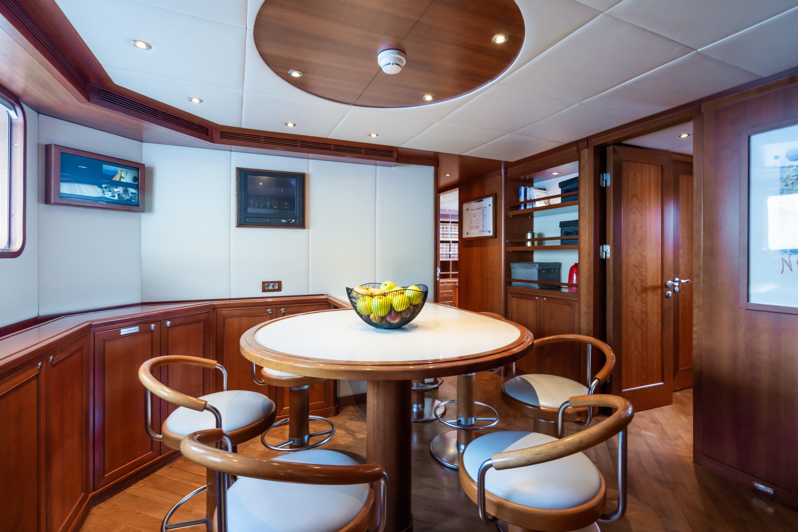 A modern yacht dining area with wood paneling, a round table with six chairs, and a bowl of fruit in the center. The room features large windows, cabinets, and soft ceiling lights.