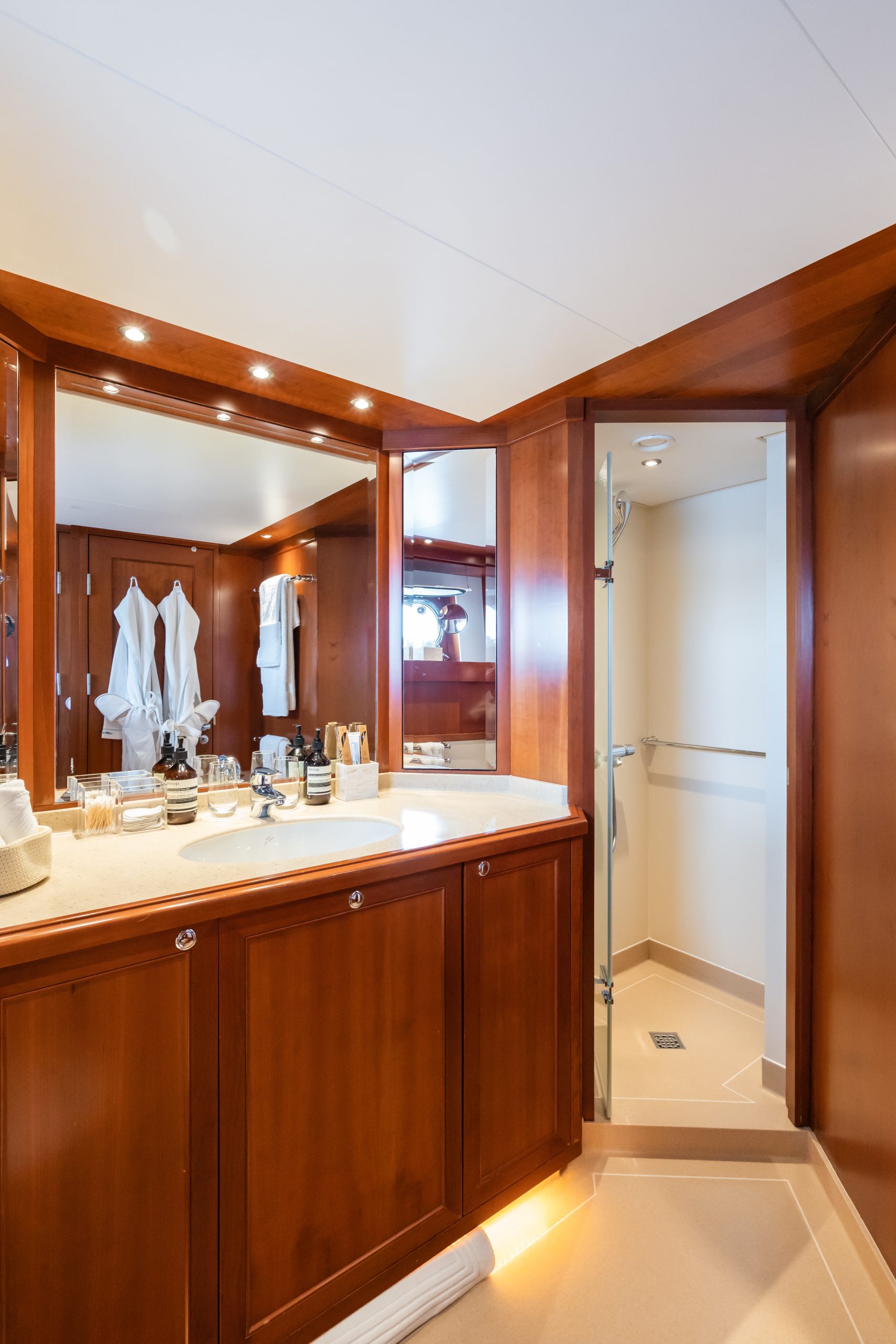 A luxurious bathroom with wooden cabinets, a wide counter with toiletries, large mirrors, white bathrobes hanging on the wall, and a walk-in shower with a glass door.