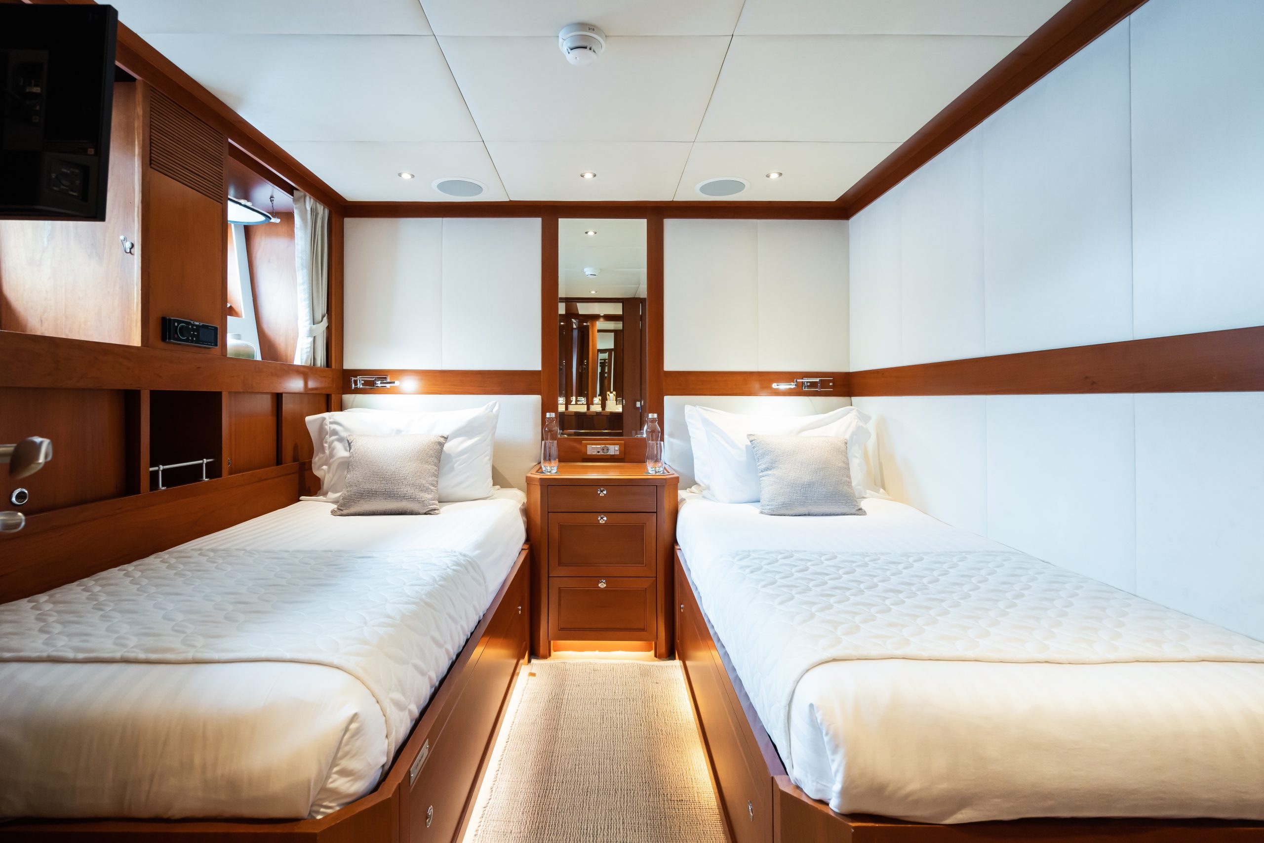 A cozy cabin with two single beds separated by a wooden nightstand, white bedding, decorative pillows, and wood panel accents. A large mirror and soft lighting create a bright and inviting atmosphere.