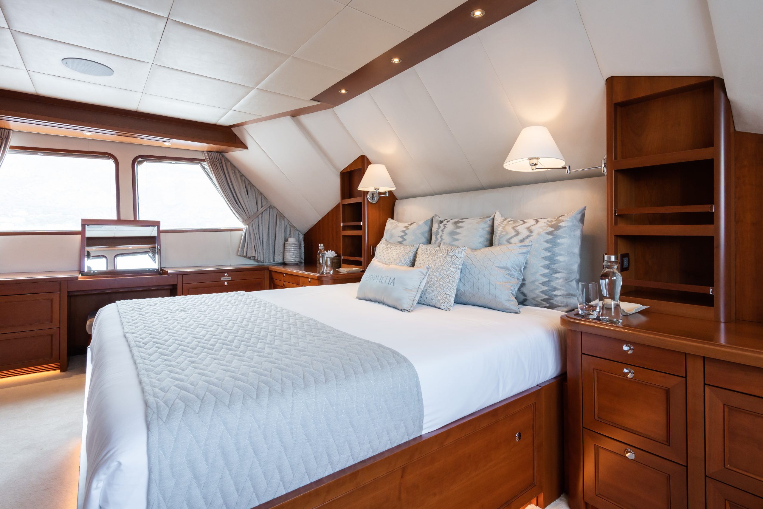 A luxurious yacht bedroom with a large bed, light blue and white bedding, wooden furniture, built-in shelves, bedside lamps, and large windows letting in natural light. Two glasses and a water bottle sit on the nightstand.