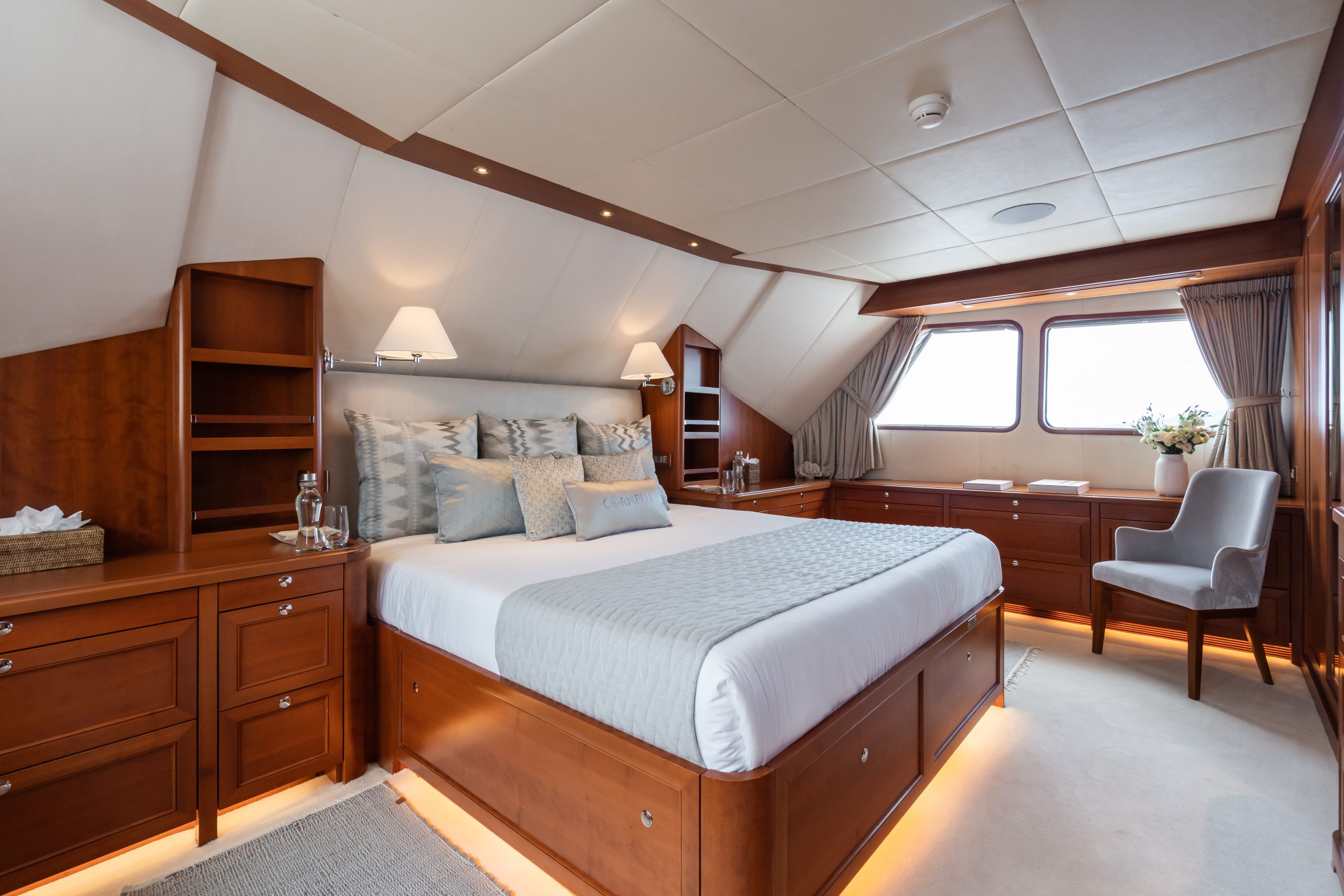 Luxurious yacht bedroom with a large bed, elegant wood furnishings, soft lighting, two windows, a vanity with a chair, decorative pillows, and neatly arranged accessories, creating a cozy and upscale atmosphere.
