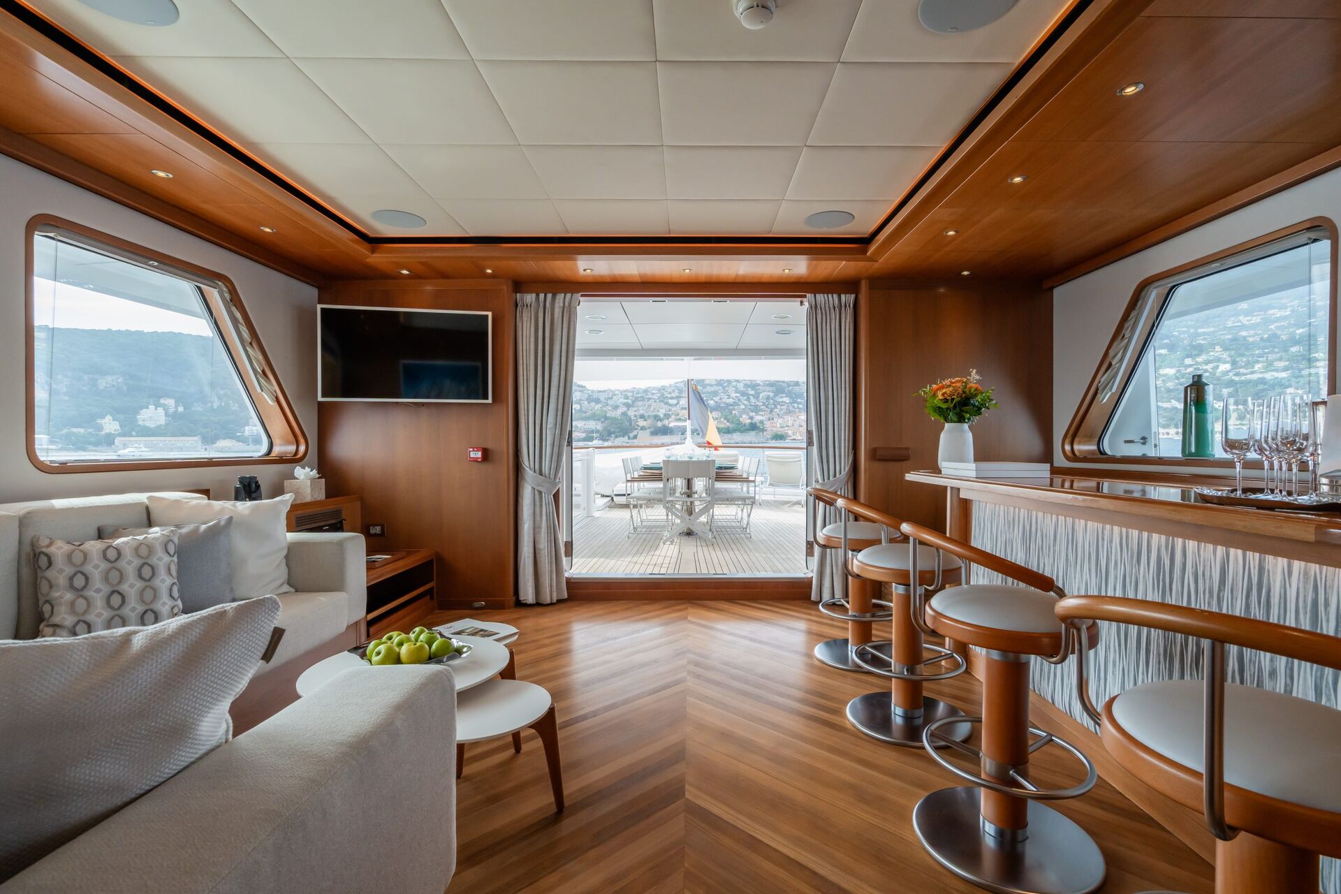 Luxurious yacht interior with wood accents, a bar with stools, sofas, a TV, and large windows. Glass doors open to an outdoor deck with a dining table and scenic coastal views in the background.