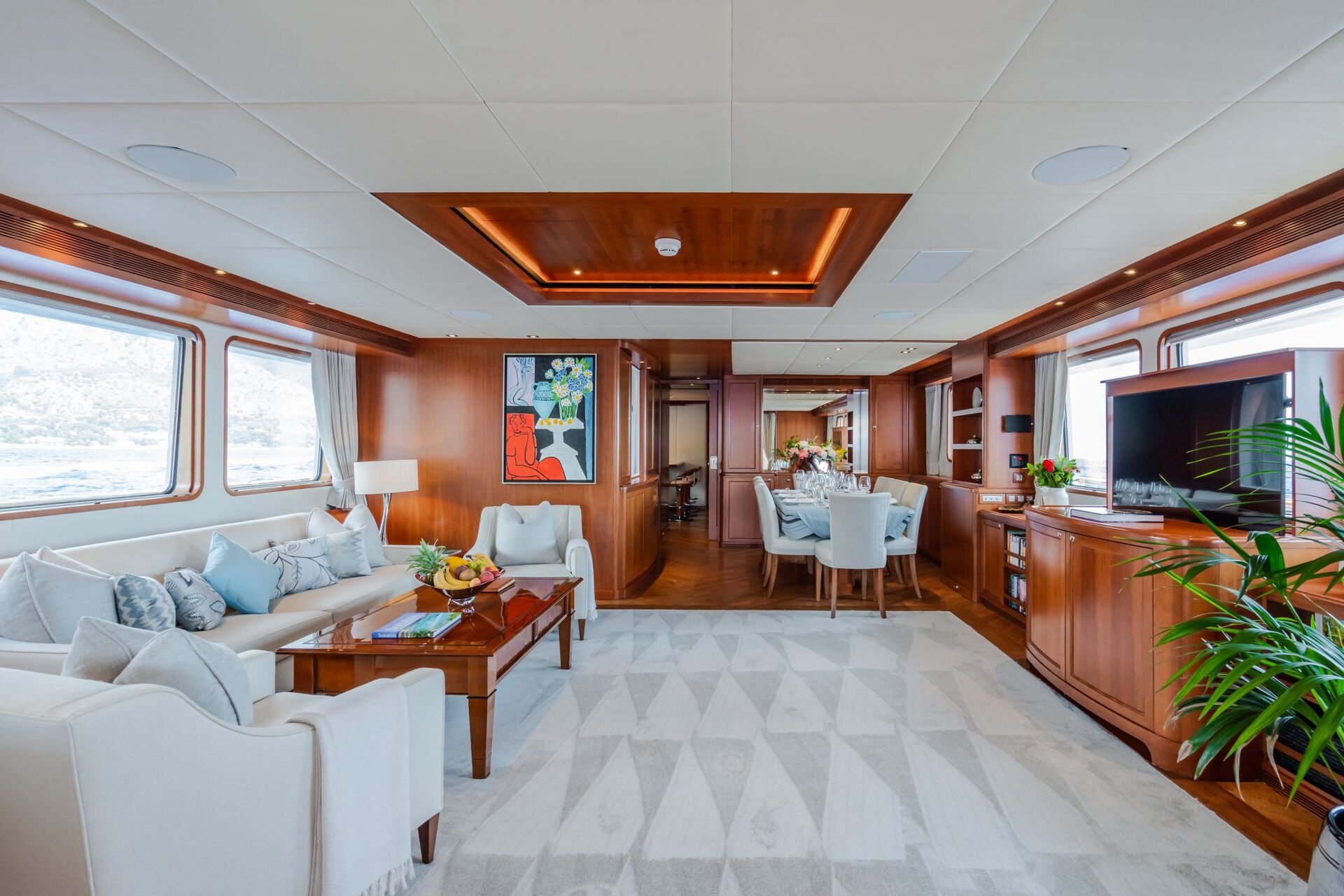 Luxurious yacht interior with wood paneling, a cream sofa set, a modern rug, large windows, a dining area with light blue chairs, wall art, a TV, plants, and views of water and mountains outside.