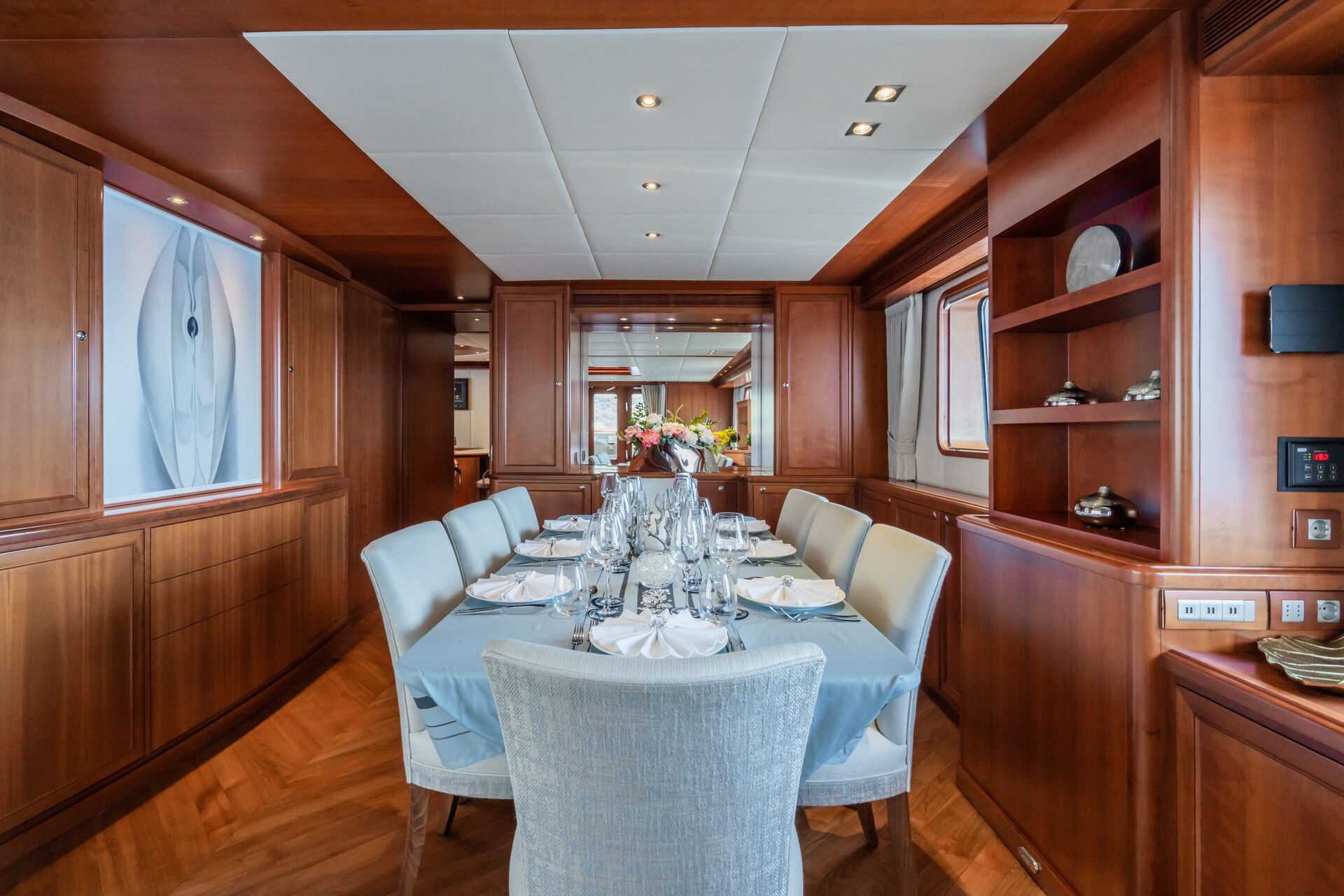 Elegant yacht dining room with a table set for eight, featuring light blue chairs, neatly arranged tableware, wood-paneled walls, shelves, and a large window letting in natural light.