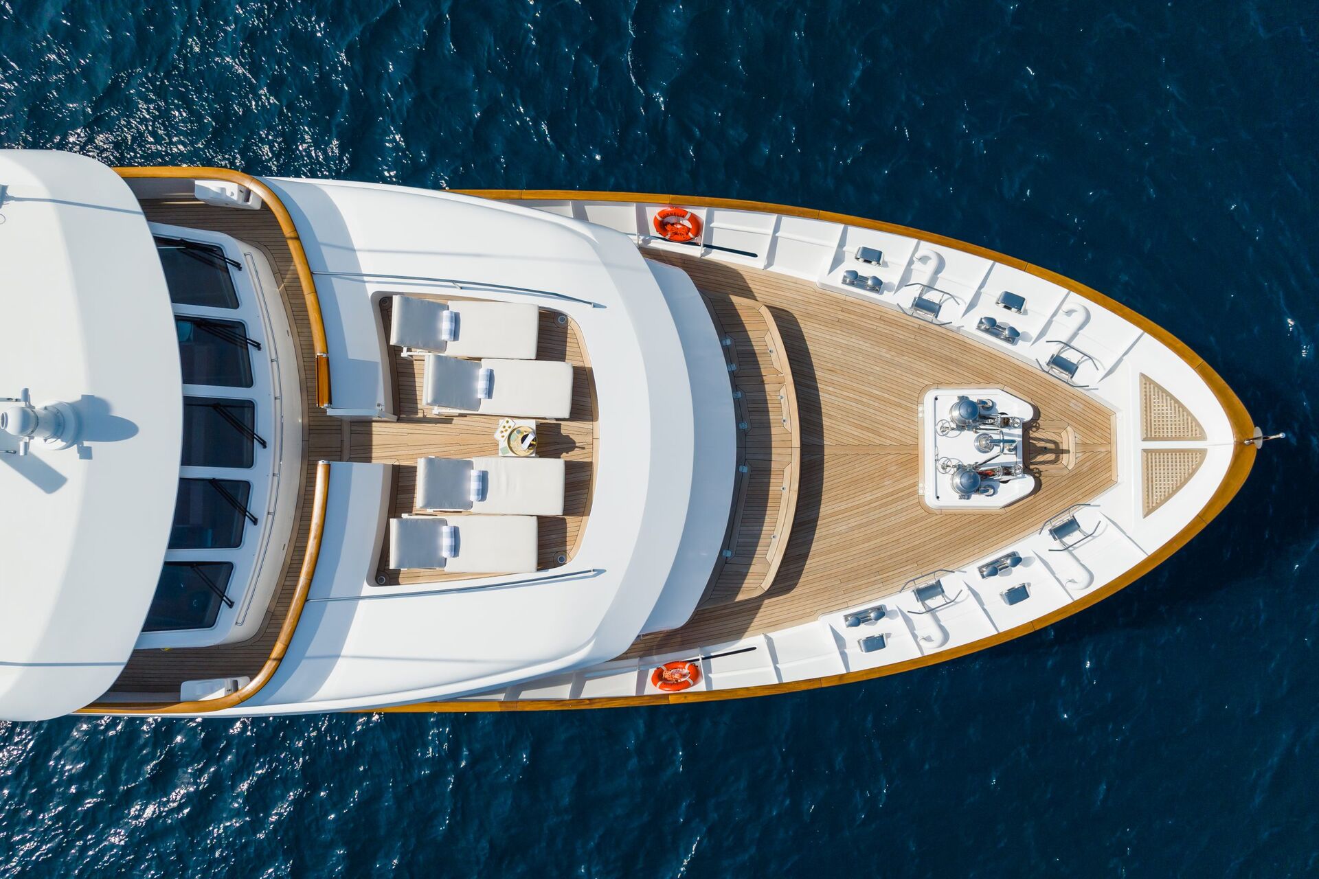 Aerial view of the bow of a luxury yacht with wooden deck, several white lounge chairs, safety rings, and clear blue water surrounding the vessel.