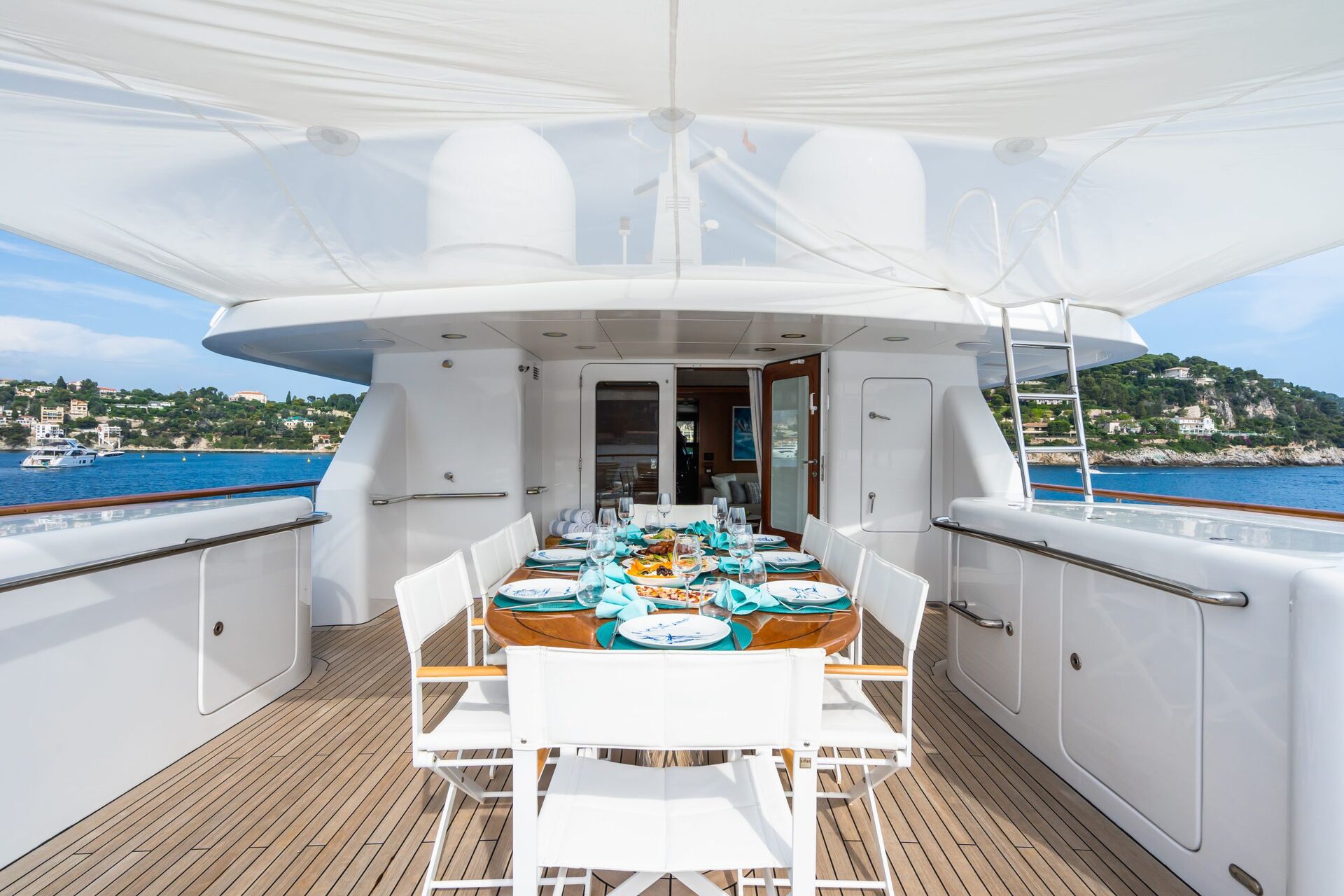 A luxurious yacht deck set for an outdoor meal, featuring a wooden table with white chairs, blue and white tableware, and a canopy overhead. The yacht is docked near a scenic coastline with houses and trees.
