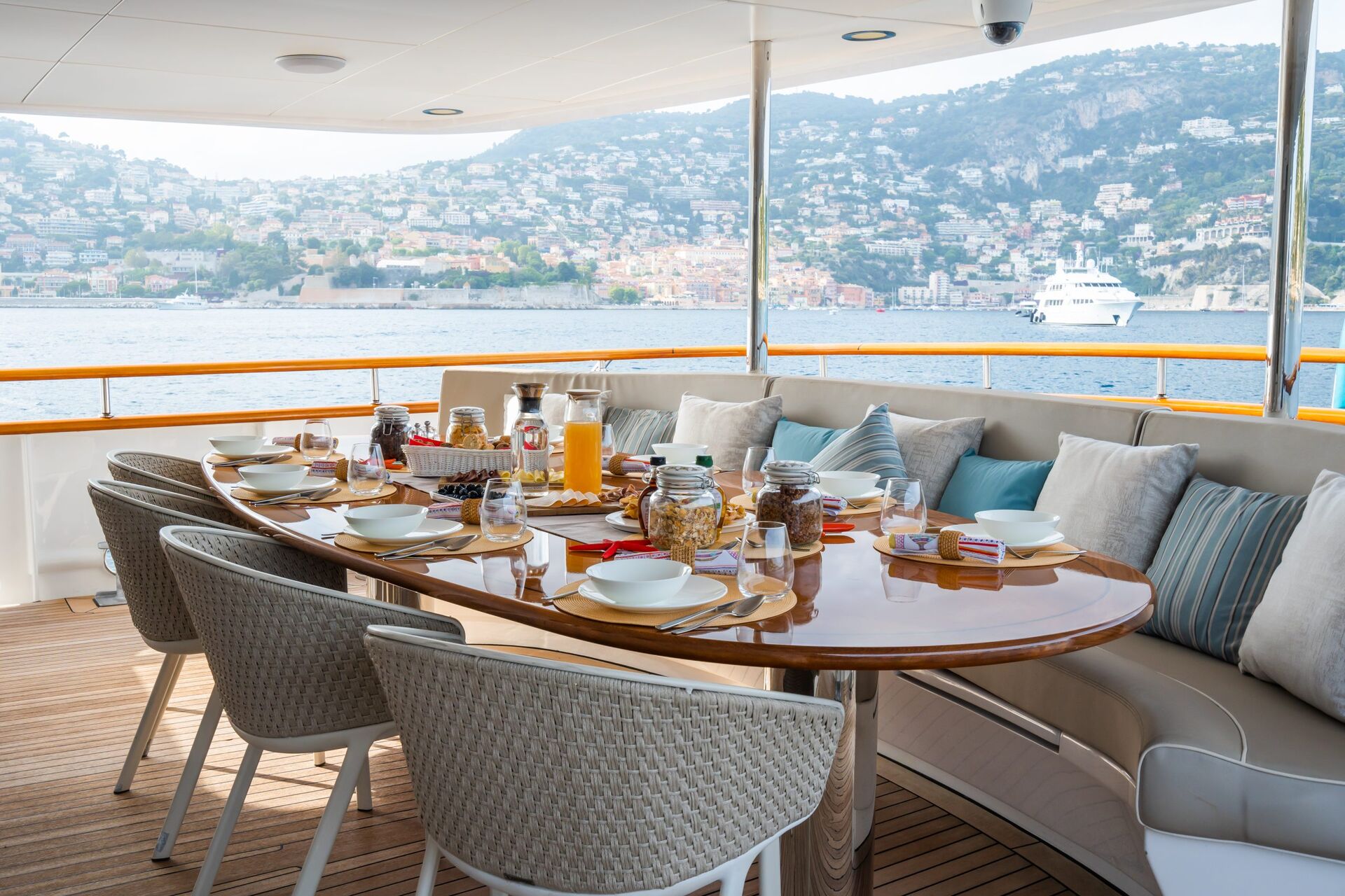 A table set for breakfast with dishes, drinks, and snacks on a yacht deck, overlooking a calm sea and a coastal town with hills and houses in the background. Cushioned seats and pillows surround the table.