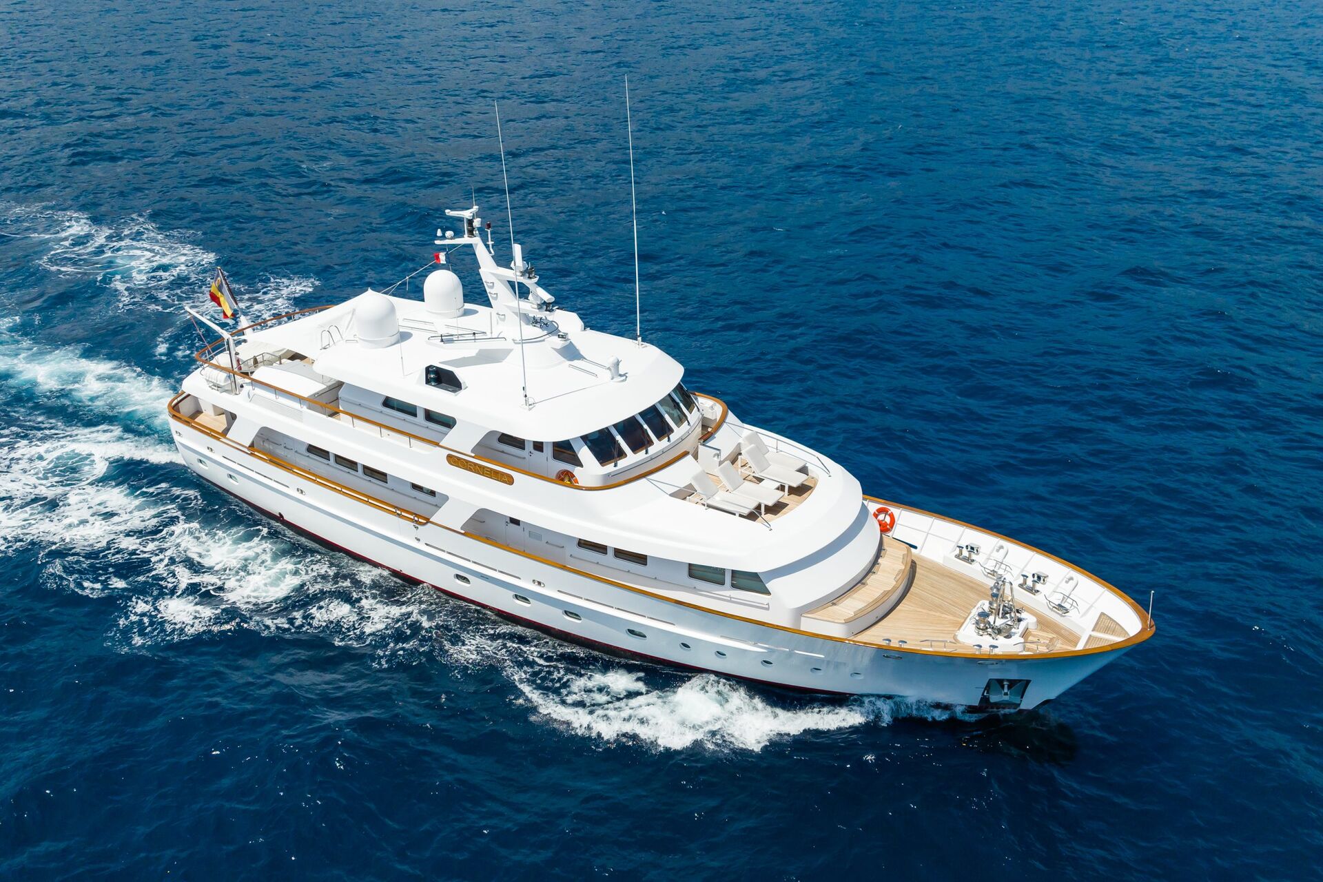 A large white yacht cruises through deep blue ocean water, leaving a foamy wake behind. The yacht has multiple decks, wooden trim, and flags flying at the stern.