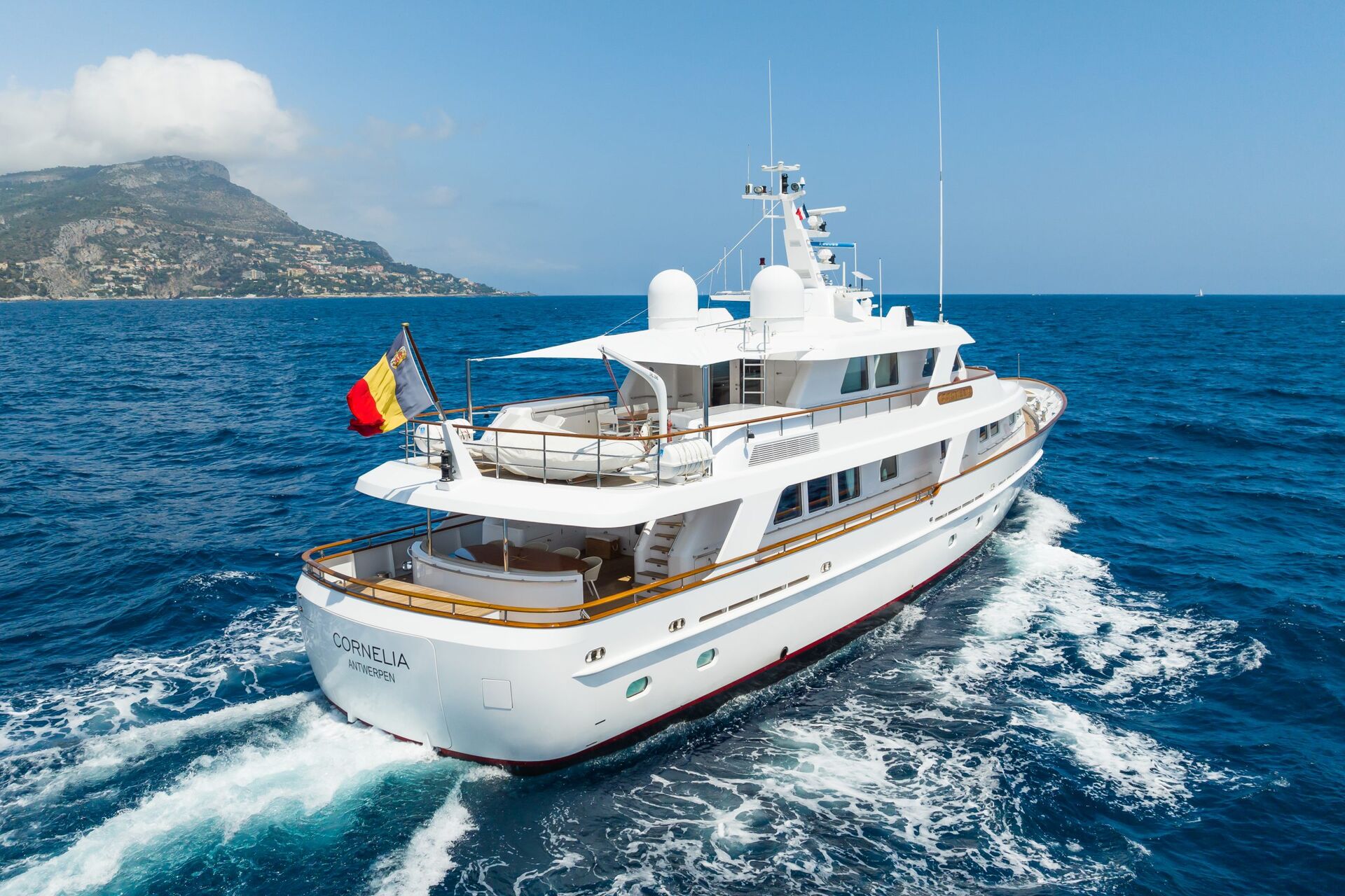 A white yacht named “Lorenilla” sails on blue ocean water near a coastline with mountains, leaving a foamy trail behind. The yacht displays a Belgian flag and is bathed in sunlight under a clear sky.