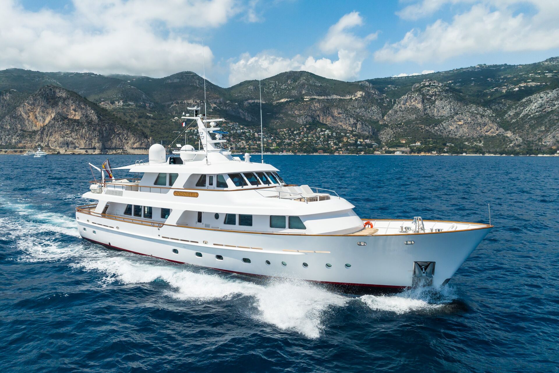 A large white luxury yacht cruises through deep blue water with mountains and green hills in the background under a partly cloudy sky.