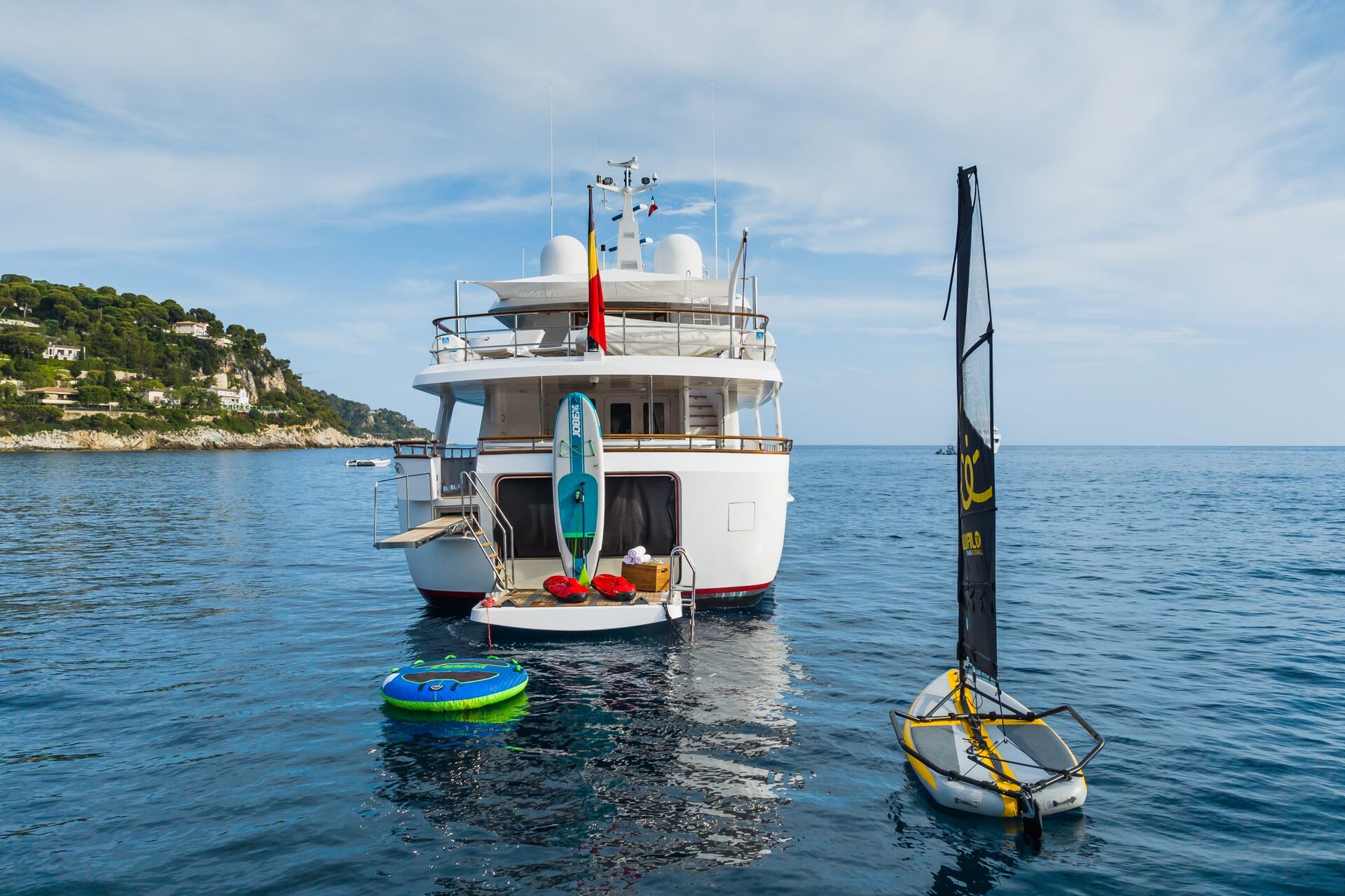 A white yacht is anchored in calm blue water, with an inflatable ring, paddleboard, and small sailboat nearby. Hills with houses and greenery are visible along the coastline under a partly cloudy sky.