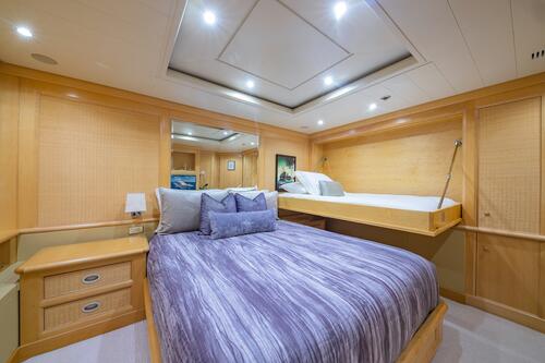 A cozy bedroom with wood paneling features a double bed with purple bedding, a pull-down bunk bed above, nightstands with lamps, and a large mirror reflecting part of the room.