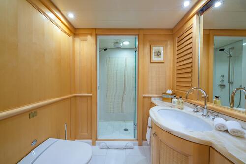 A well-lit bathroom with light wood paneling, a glass-enclosed shower, a white toilet, a marble countertop with a dual sink, a large mirror, and neatly arranged toiletries and towels.