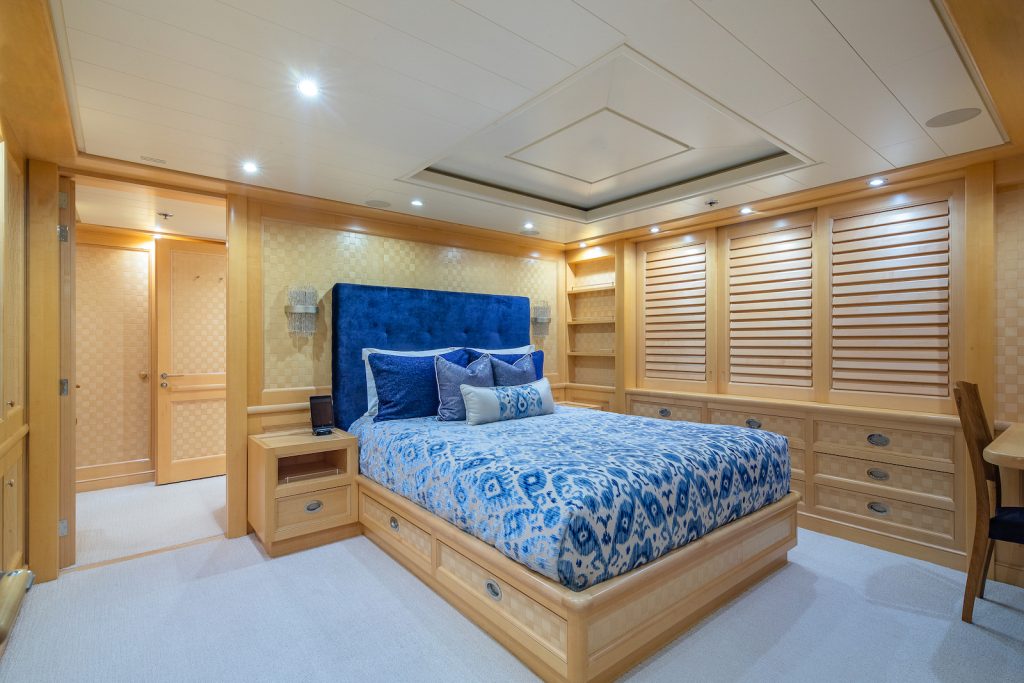 A spacious bedroom with light wood walls and furniture, a large bed with blue bedding and multiple pillows, built-in shelves, and a window with wooden shutters. Recessed ceiling lights illuminate the room.