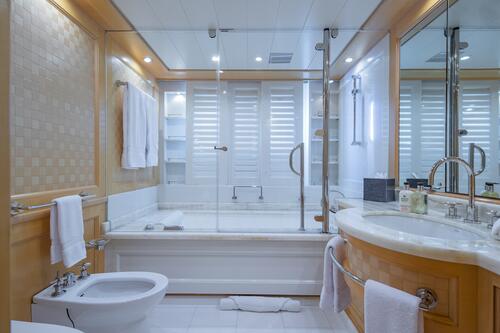 Luxurious bathroom with a large bathtub, glass shower enclosure, bidet, sink with curved countertop, towels, and toiletries. White shutters cover the windows, and the room has warm wood and white accents.