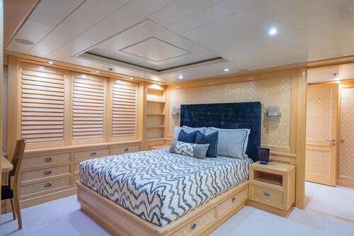 A modern bedroom with wood paneling, built-in shelves, a desk, and a large bed featuring a blue headboard and chevron bedding. Recessed lighting and storage drawers are visible under the bed and along the wall.